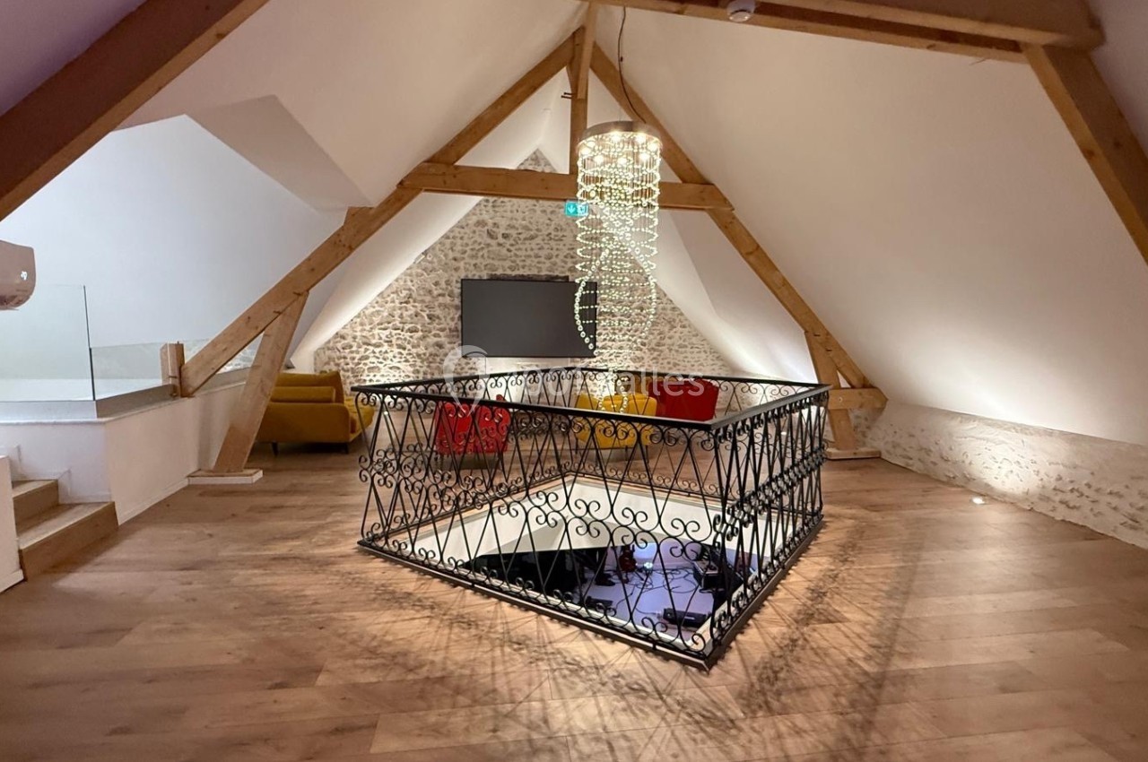 Grenier aménagé avec parquet, murs en pierre, poutres apparentes, escalier central entouré d'une rambarde en fer forgé.