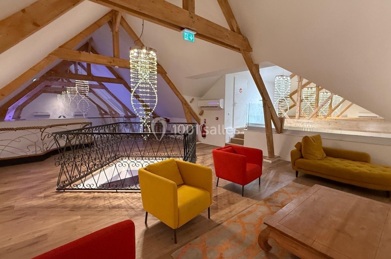 Espace mansardé avec poutres apparentes, fauteuils colorés, table basse en bois et lustres modernes suspendus.