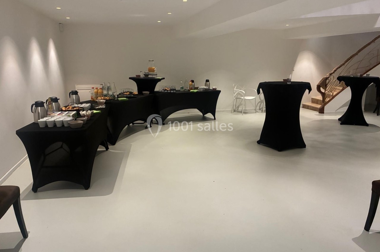 Salle lumineuse avec tables noires dressées pour un buffet, comprenant boissons, collations et chaises en arrière-plan.