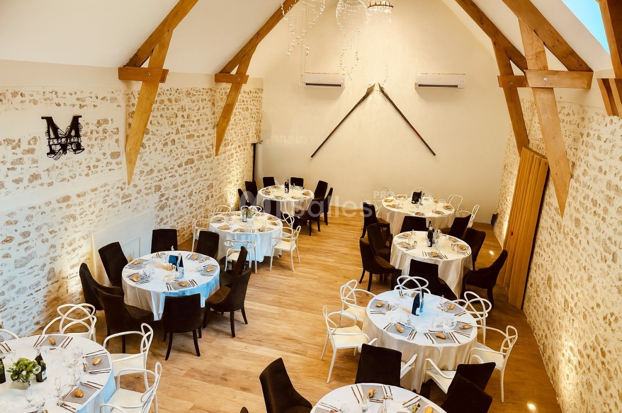 Salle de réception lumineuse avec murs en pierre, poutres apparentes et tables rondes dressées pour un événement.