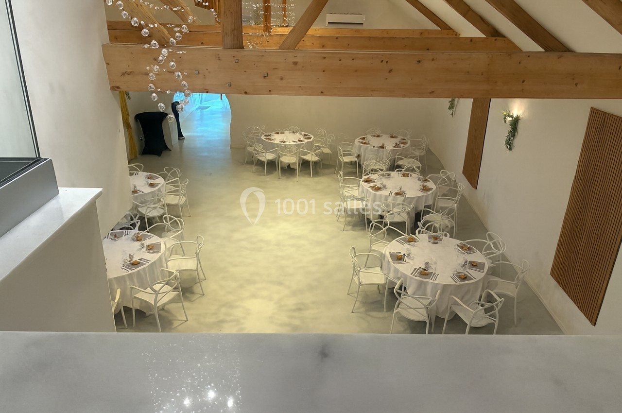 Salle lumineuse avec tables rondes dressées, chaises blanches et poutres apparentes en bois.
