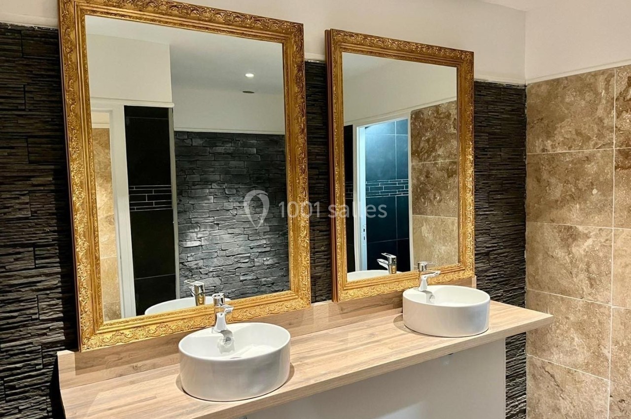 Deux lavabos blancs avec miroirs dorés sur un plan en bois, entourés de murs en pierre et carrelage beige.