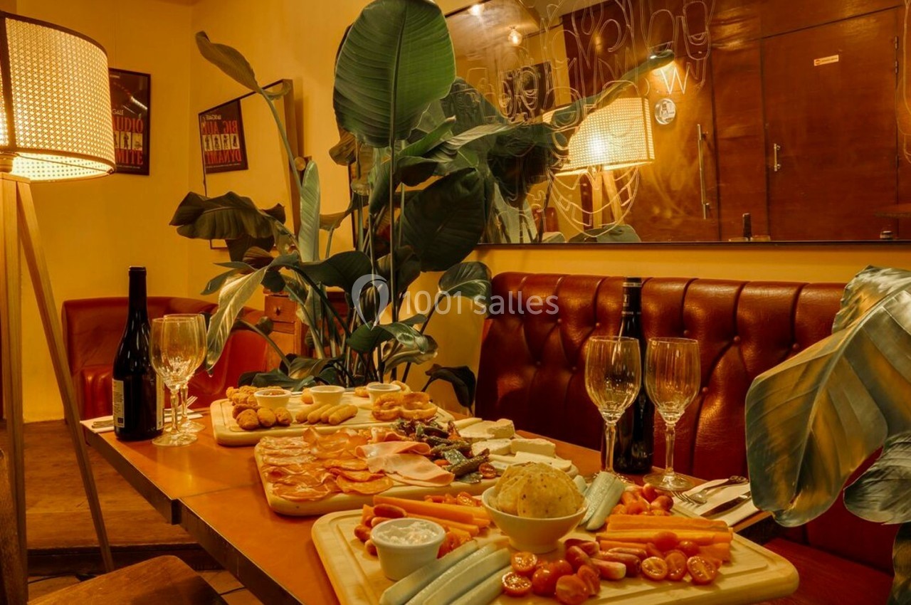 Table dressée dans un restaurant chaleureux avec planches de charcuterie, légumes, fromages, vin rouge et décor végétal.