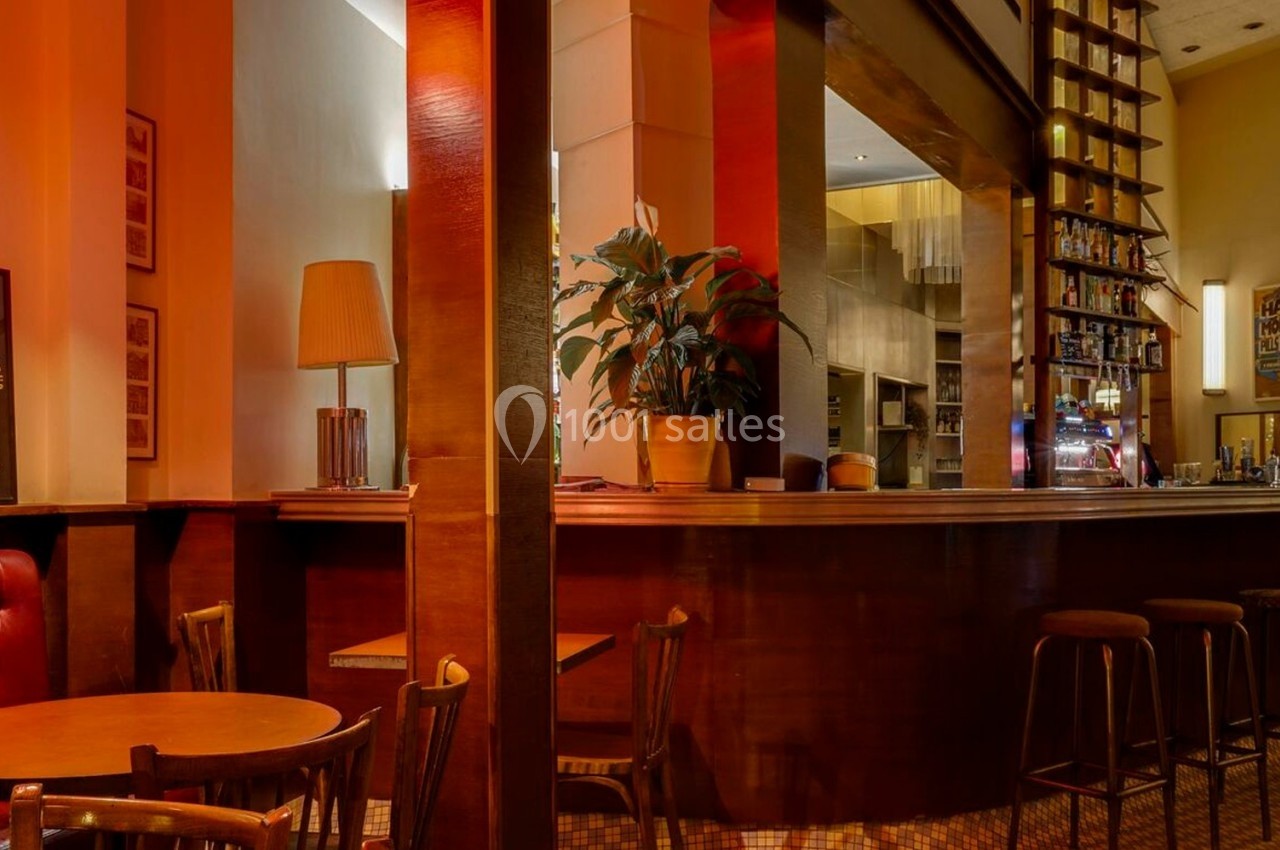 Intérieur d'un café chaleureux avec comptoir en bois, tabourets hauts, tables rondes et éclairage tamisé.