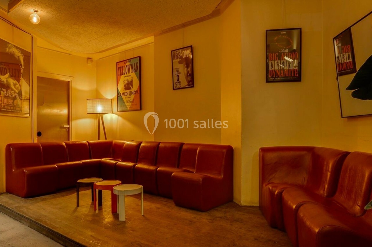 Salon avec canapés en cuir rouge, tables basses colorées et affiches encadrées sur des murs jaunes.