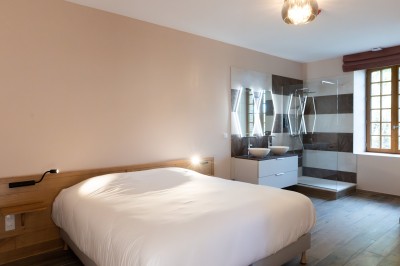 Chambre avec lit double, poutres apparentes, linge de lit blanc, et salle de bain attenante visible par une porte ouverte.