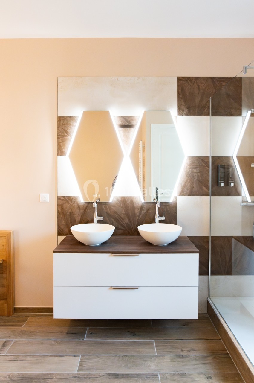 Double vasque moderne avec miroir rétroéclairé en forme hexagonale, carrelage beige et sol en bois clair.