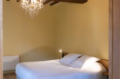 Chambre avec lit double, poutres apparentes, linge de lit blanc, et salle de bain attenante visible par une porte ouverte.