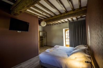 Chambre avec lit double, poutres apparentes, linge de lit blanc, et salle de bain attenante visible par une porte ouverte.