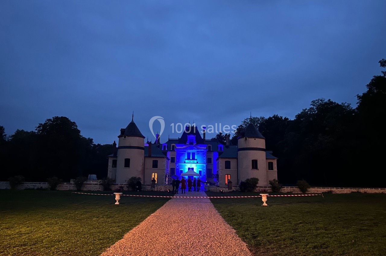 Façade d'un château éclairée en bleu à la tombée de la nuit, entourée de verdure et d'un chemin gravillonné.