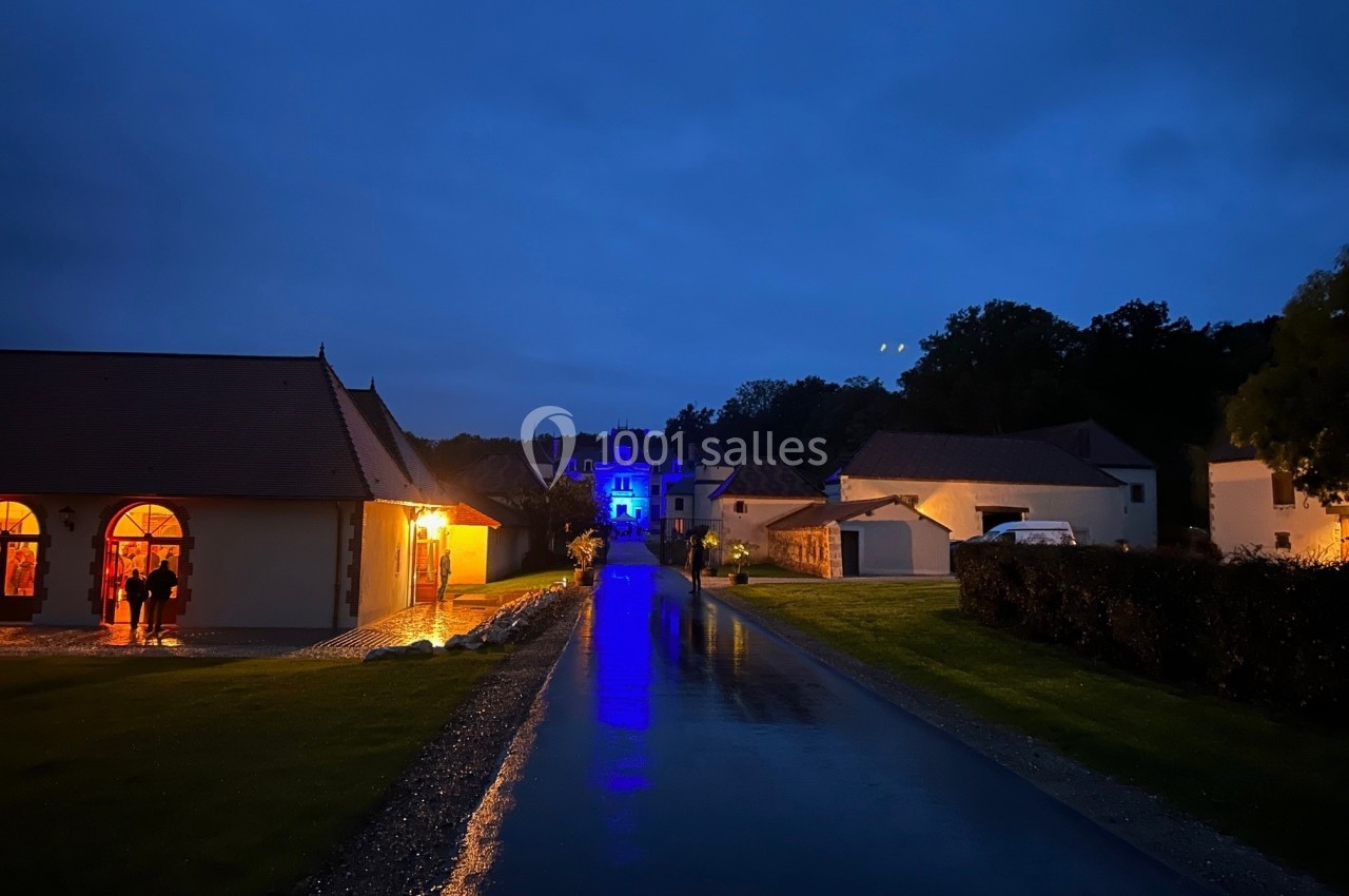 Chemin pavé éclairé menant à un bâtiment illuminé en bleu, entouré de maisons dans un cadre nocturne.