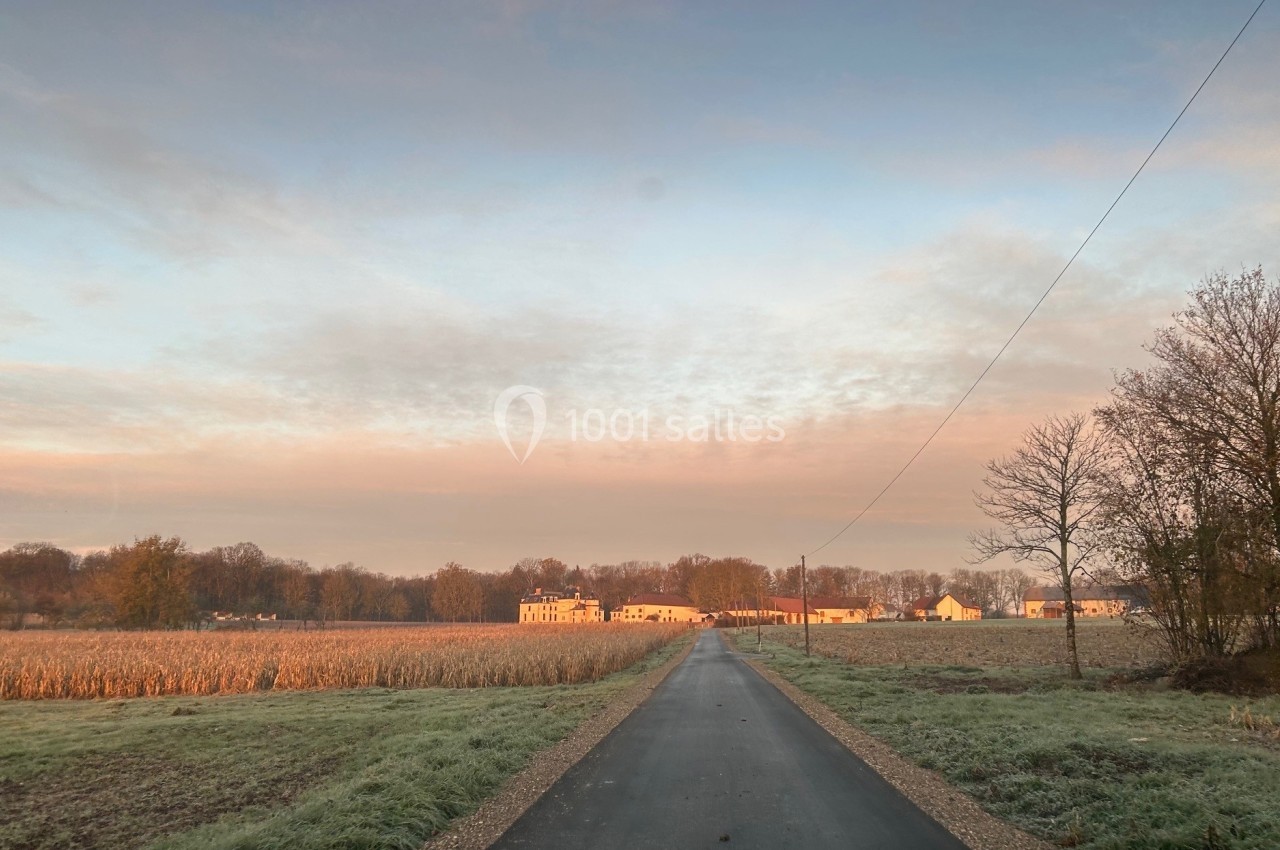 Route étroite traversant une campagne avec champs, arbres et bâtiments au loin sous un ciel partiellement nuageux.