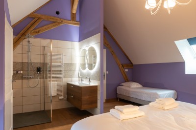 Chambre avec lit double, poutres apparentes, linge de lit blanc, et salle de bain attenante visible par une porte ouverte.