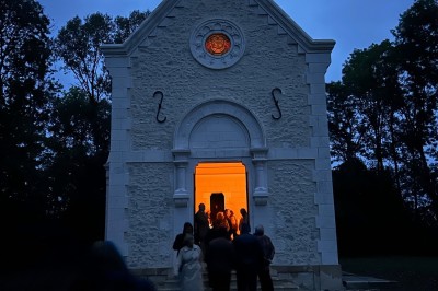 Chambre avec lit double, poutres apparentes, linge de lit blanc, et salle de bain attenante visible par une porte ouverte.