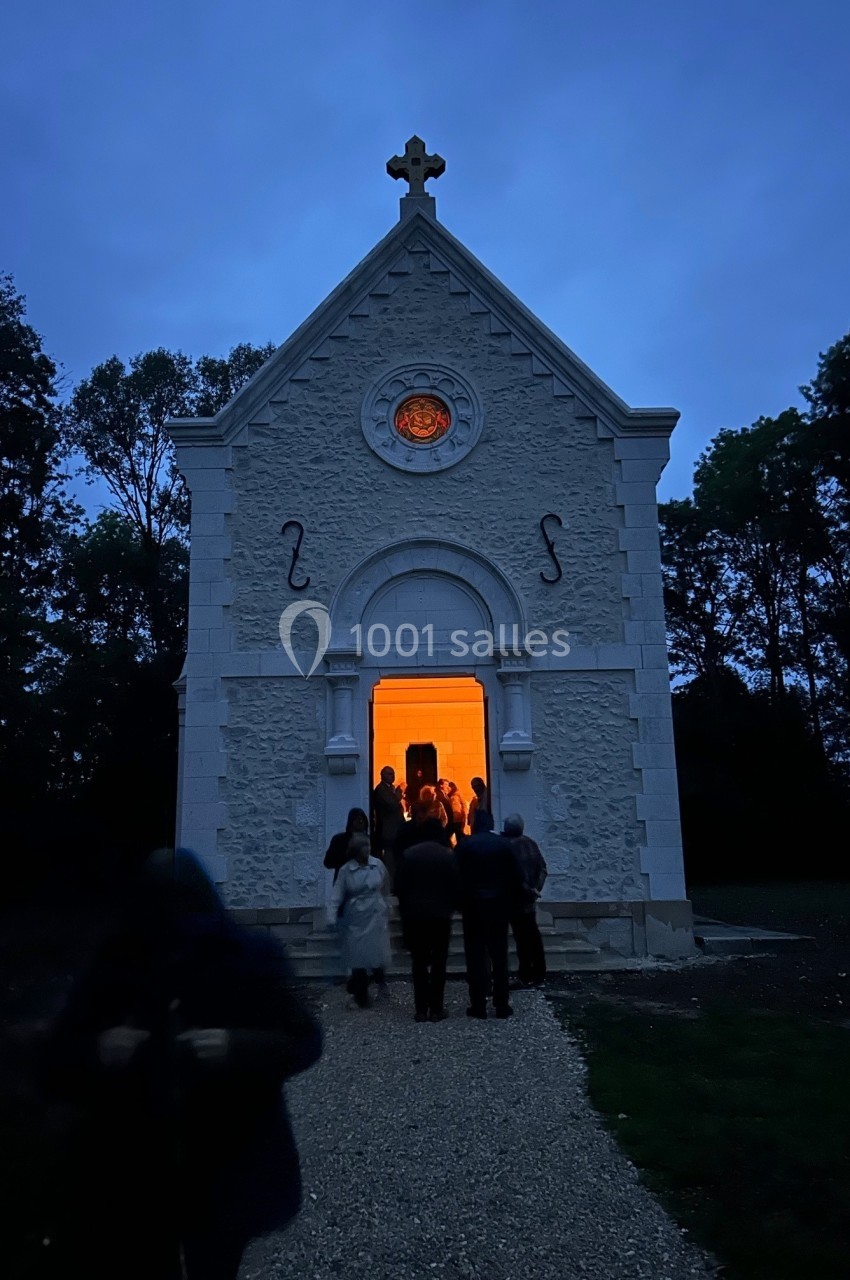 Petite chapelle blanche éclairée de l'intérieur, entourée de visiteurs à la tombée de la nuit.