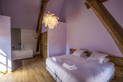 Chambre avec lit double, poutres apparentes, linge de lit blanc, et salle de bain attenante visible par une porte ouverte.