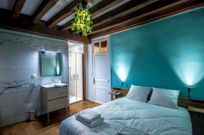 Chambre avec lit double, poutres apparentes, linge de lit blanc, et salle de bain attenante visible par une porte ouverte.