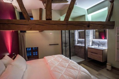 Chambre avec lit double, poutres apparentes, linge de lit blanc, et salle de bain attenante visible par une porte ouverte.