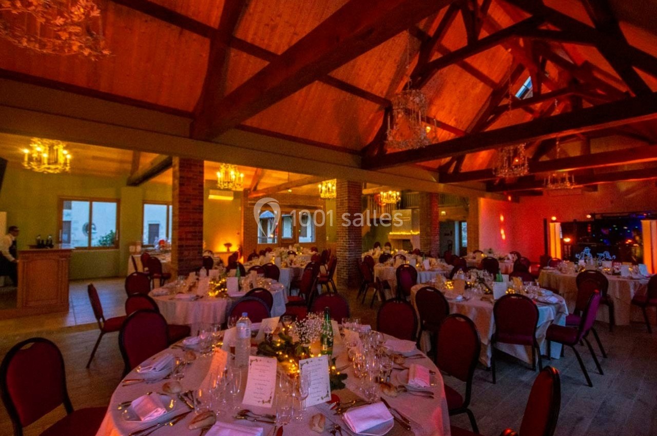 Salle de réception chaleureuse avec tables rondes dressées, chaises rouges et éclairage tamisé sous une charpente en bois.