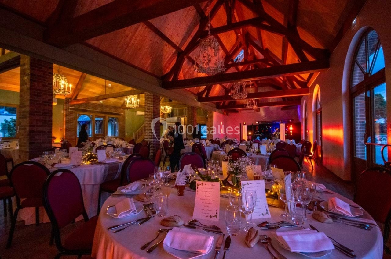 Salle de réception décorée avec des tables rondes dressées, éclairage chaleureux et poutres apparentes au plafond.