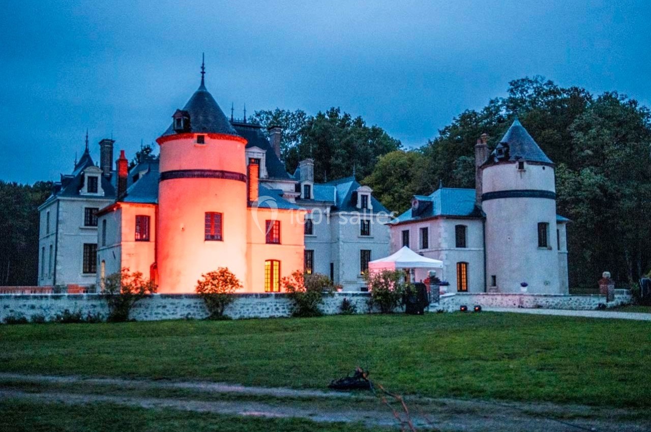 Château éclairé par des lumières rouges au crépuscule, entouré de pelouses et d'arbres.