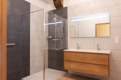 Chambre avec lit double, poutres apparentes, linge de lit blanc, et salle de bain attenante visible par une porte ouverte.