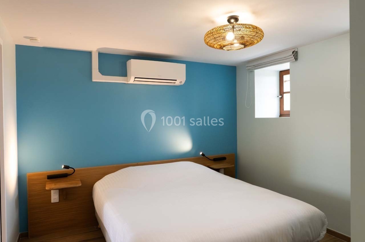 Chambre avec un lit double, mur bleu, éclairage suspendu, climatisation murale et petite fenêtre avec store.