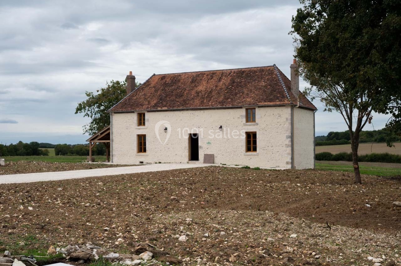 Maison en pierre blanche avec toit en tuiles rouges, entourée d'un terrain dégagé et d'arbres sous un ciel nuageux.