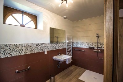 Chambre avec lit double, poutres apparentes, linge de lit blanc, et salle de bain attenante visible par une porte ouverte.