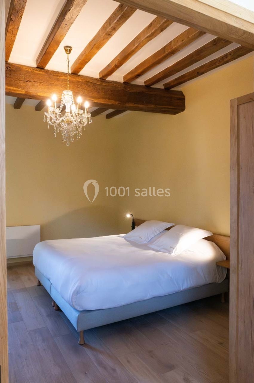 Chambre avec lit double, parquet clair, murs jaunes, poutres apparentes et lustre en cristal.