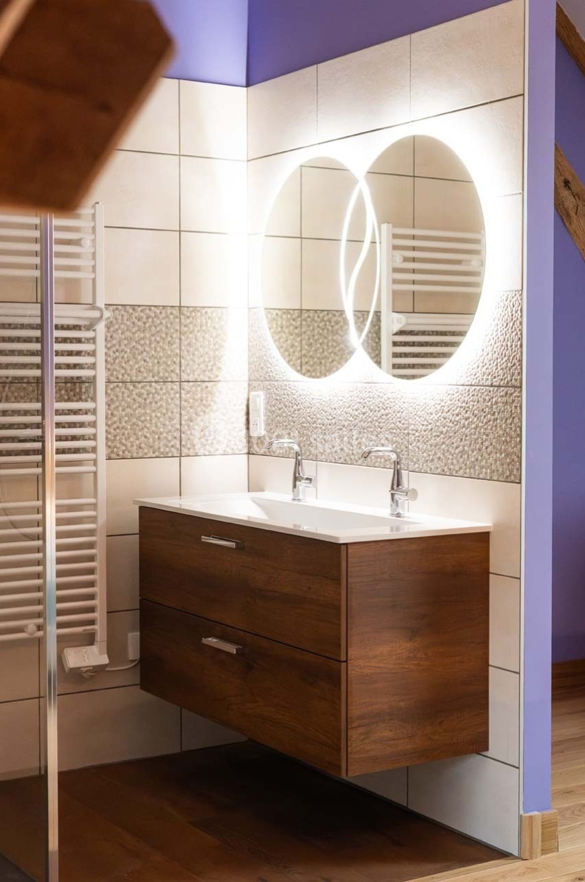 Meuble de salle de bain en bois avec double vasque, miroir rond rétroéclairé et carrelage mural texturé.