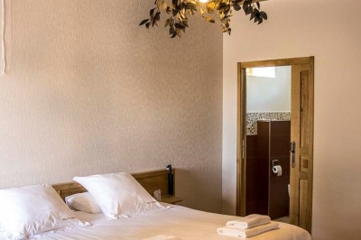 Chambre avec lit double, poutres apparentes, linge de lit blanc, et salle de bain attenante visible par une porte ouverte.