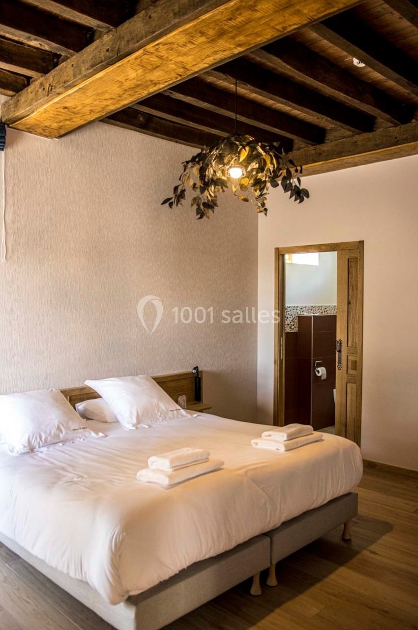 Chambre avec lit double, poutres apparentes, linge de lit blanc, et salle de bain attenante visible par une porte ouverte.