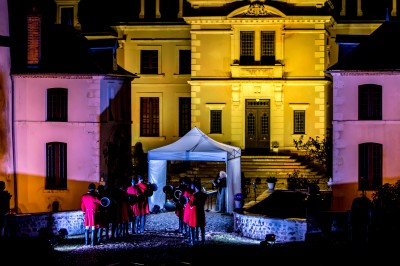 Groupe de musiciens en uniforme rouge jouant devant un bâtiment éclairé de nuit, avec une tente blanche au centre.