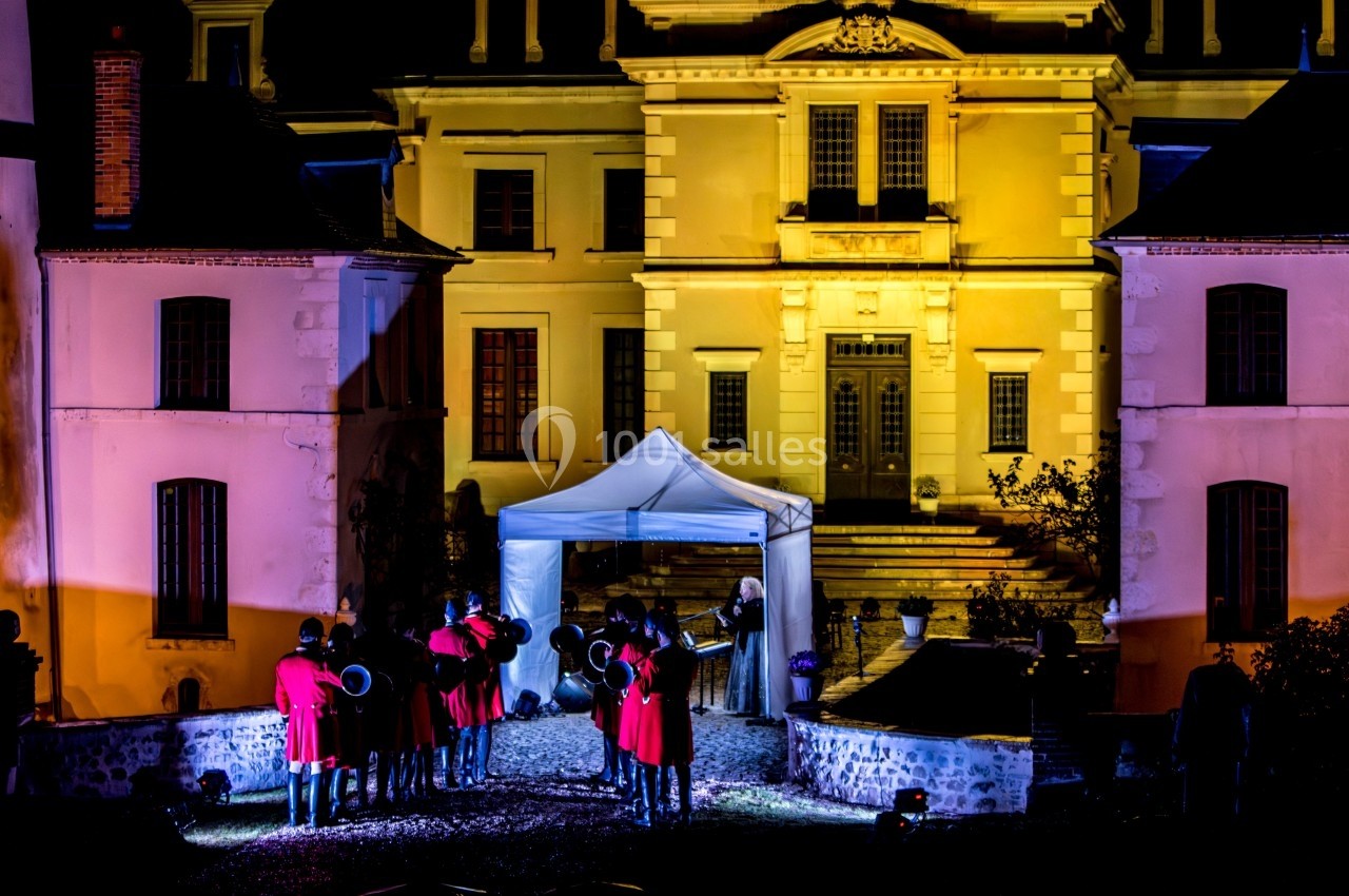 Groupe de musiciens en uniforme rouge jouant devant un bâtiment éclairé de nuit, avec une tente blanche au centre.