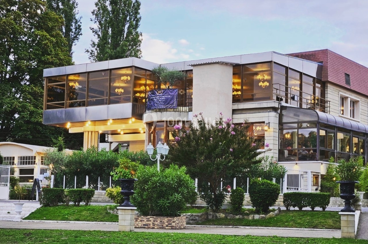 Bâtiment moderne avec de grandes baies vitrées entouré de verdure et d'arbustes, sous un ciel partiellement nuageux.