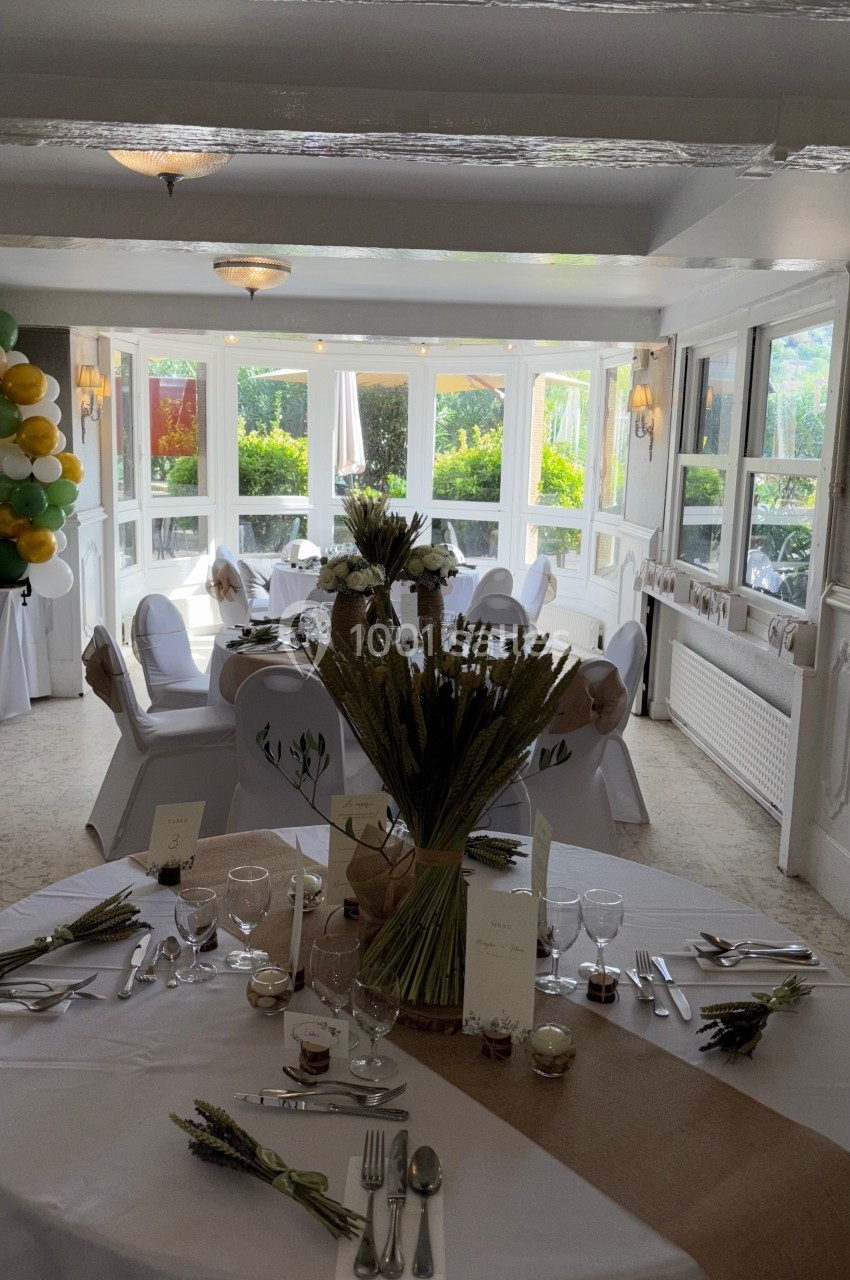 Salle de réception lumineuse décorée pour un événement, avec tables dressées, fleurs séchées et chaises ornées.