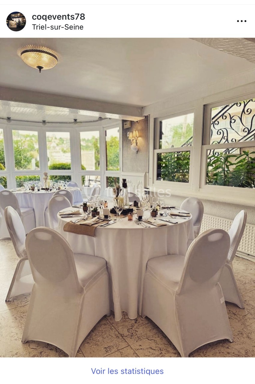 Salle lumineuse avec tables rondes dressées, nappes blanches, vaisselle élégante et vue sur un jardin verdoyant.