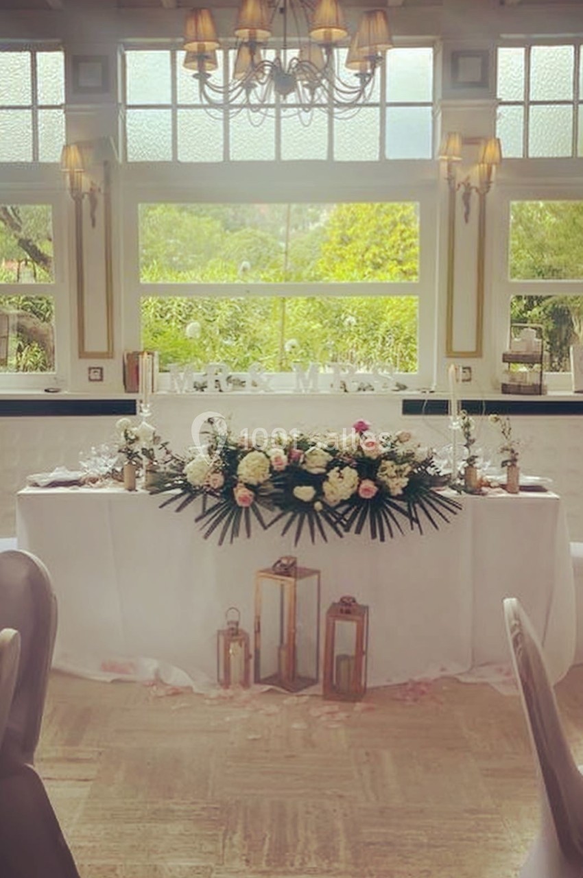 Table de mariage décorée de fleurs blanches et roses, lanternes dorées et vue sur un jardin à travers de grandes fenêtres.