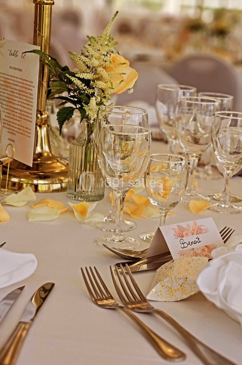 Table élégamment dressée avec vaisselle en verre, couverts dorés, fleurs jaunes et menu sur un support doré.