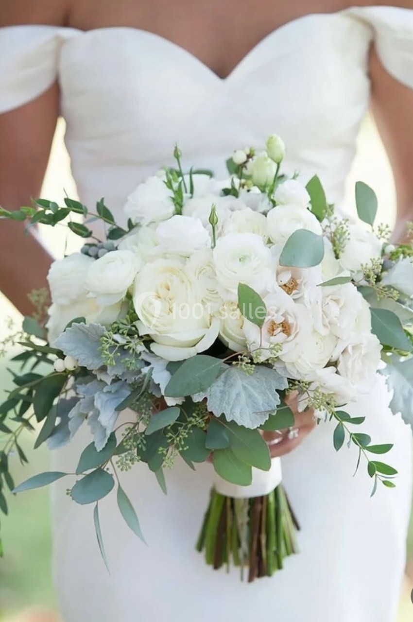 Femme en robe de mariée tenant un bouquet de fleurs blanches et feuillages verts, en extérieur.