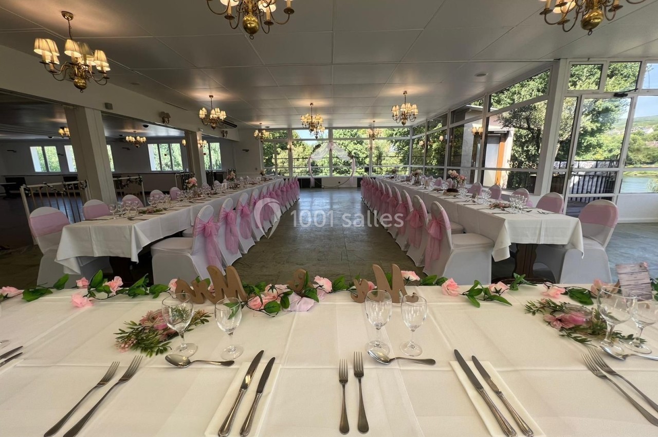 Salle de réception lumineuse avec deux longues tables dressées, décorées de fleurs roses et chaises blanches.