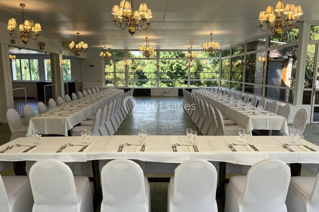 Salle de réception lumineuse avec de grandes baies vitrées, tables dressées en U et chaises blanches alignées.