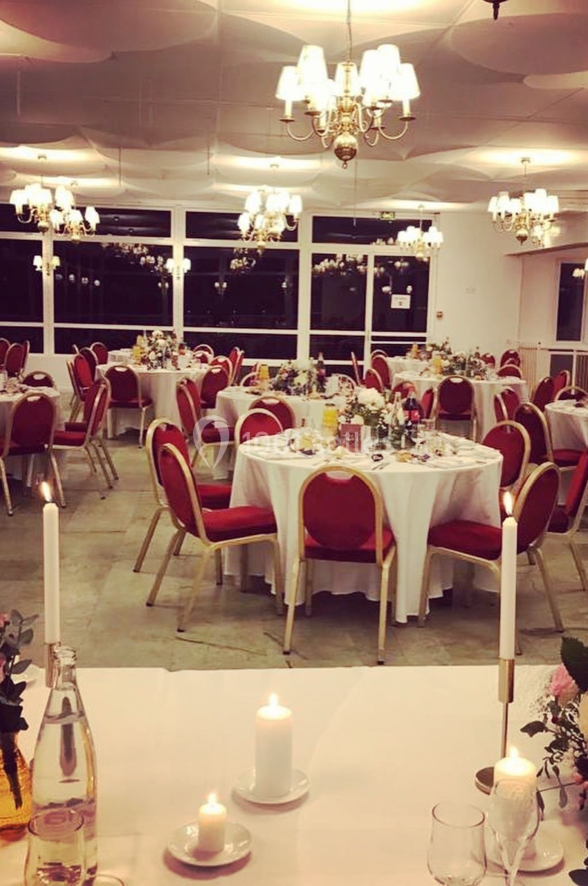 Salle de réception décorée avec des tables rondes dressées, chaises rouges et lustres suspendus.