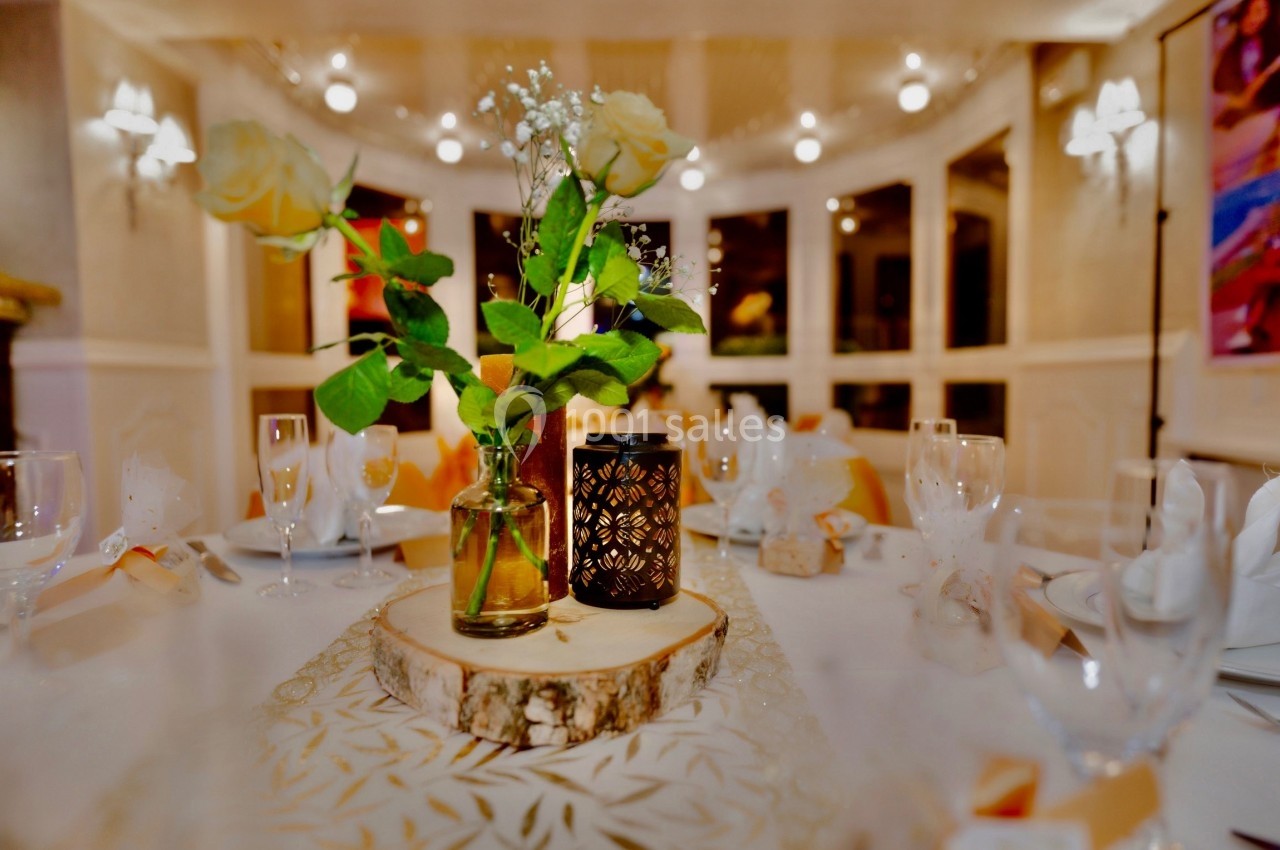 Centre de table décoratif avec roses blanches, bougie et vase sur une rondelle de bois dans une salle éclairée.