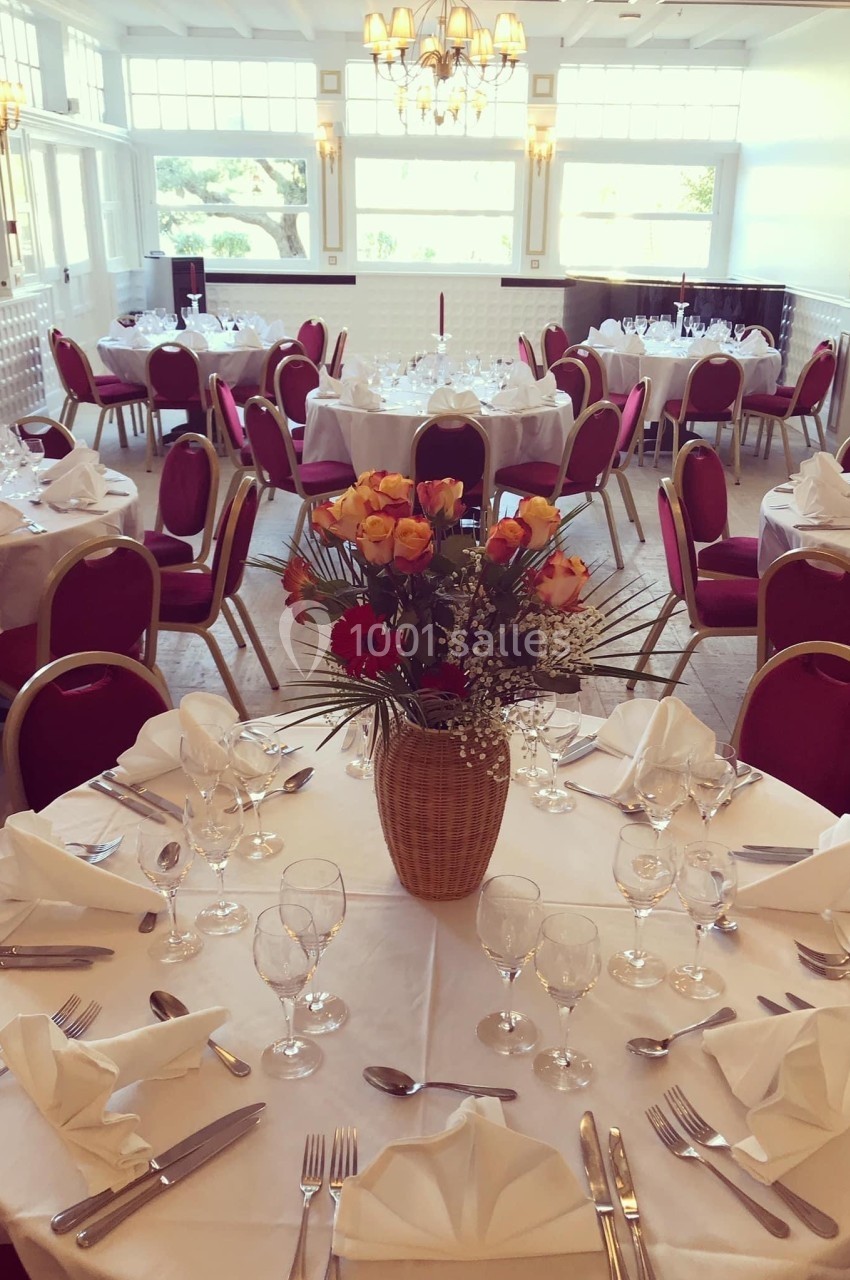 Salle de réception lumineuse avec tables rondes dressées, nappes blanches, chaises rouges et centre de table floral.