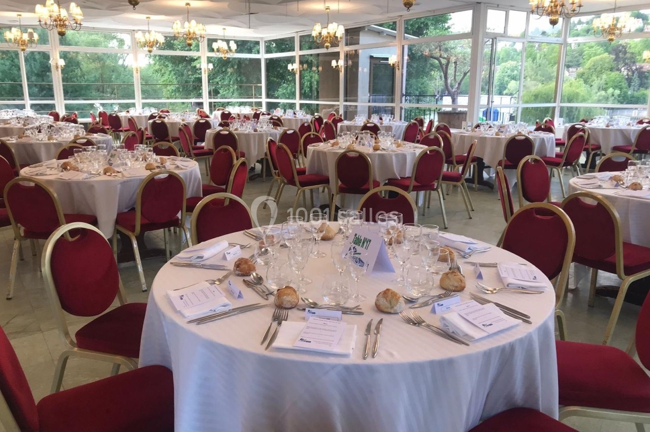 Salle de réception lumineuse avec des tables rondes dressées, nappes blanches et chaises rouges, entourée de baies vitrées.