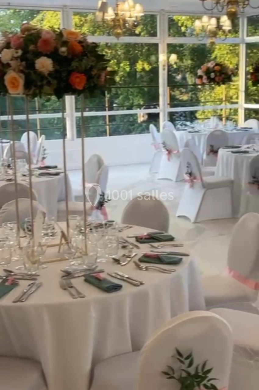 Salle de réception lumineuse avec tables rondes dressées, nappes blanches, chaises décorées et compositions florales.