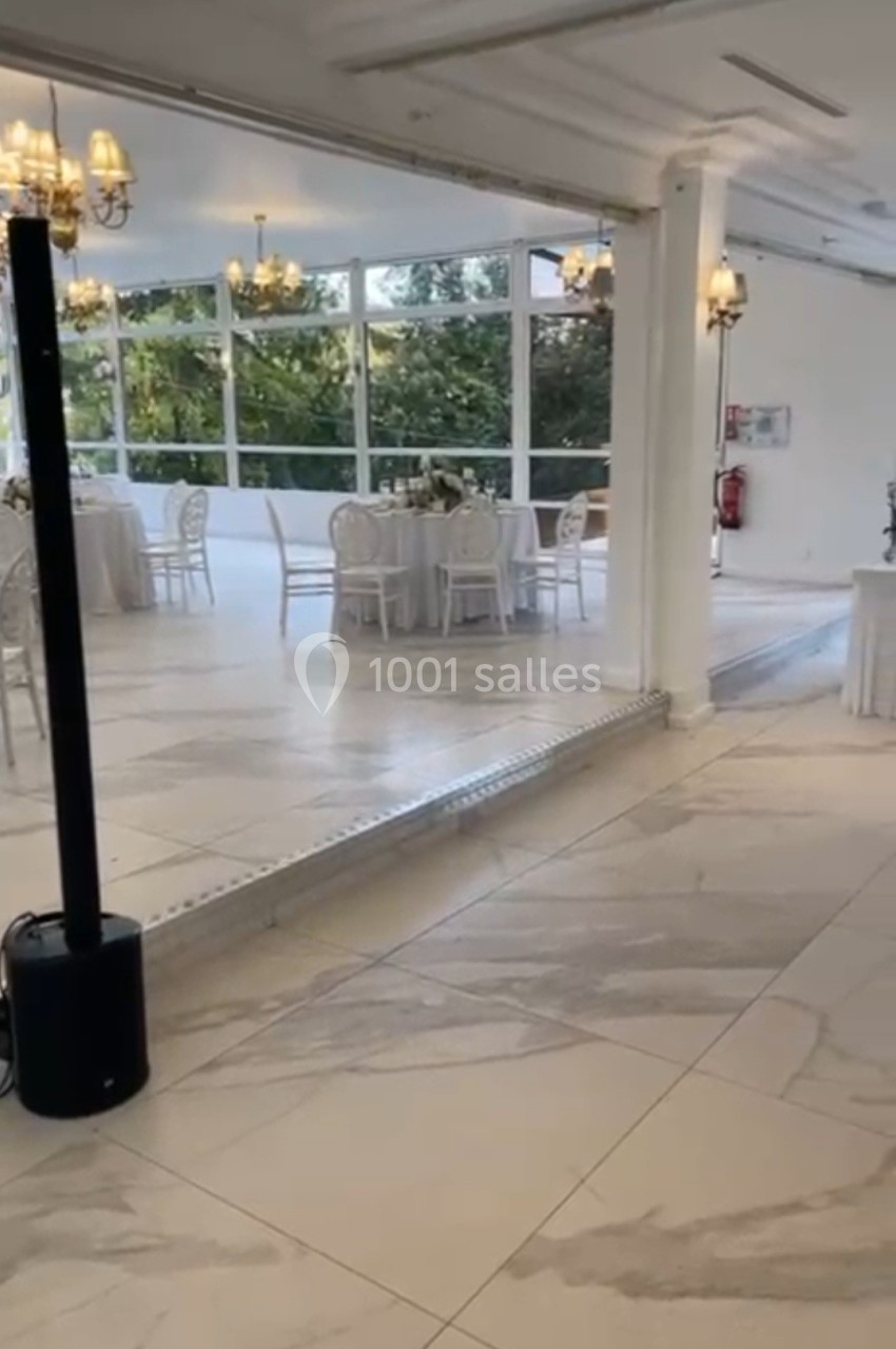 Salle lumineuse avec grandes baies vitrées, tables rondes dressées et sol en marbre clair.