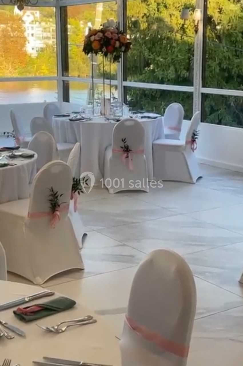 Salle de réception lumineuse avec des tables rondes dressées et des chaises décorées de rubans roses.